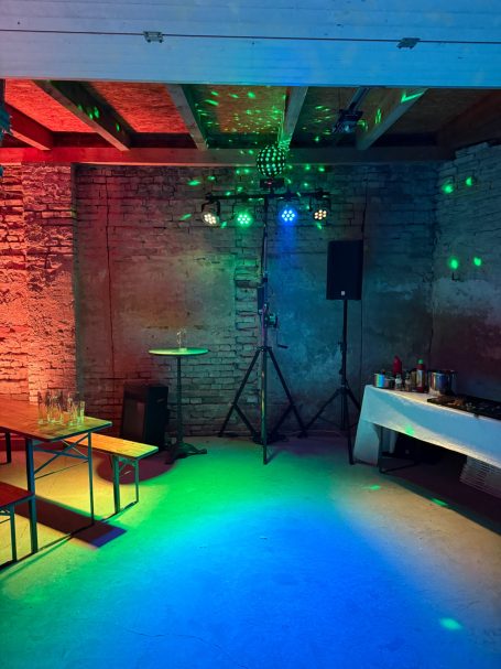 Bunte Lichtspiel von einer Party mit Stehtisch und Bänken in einem Kellerraum.